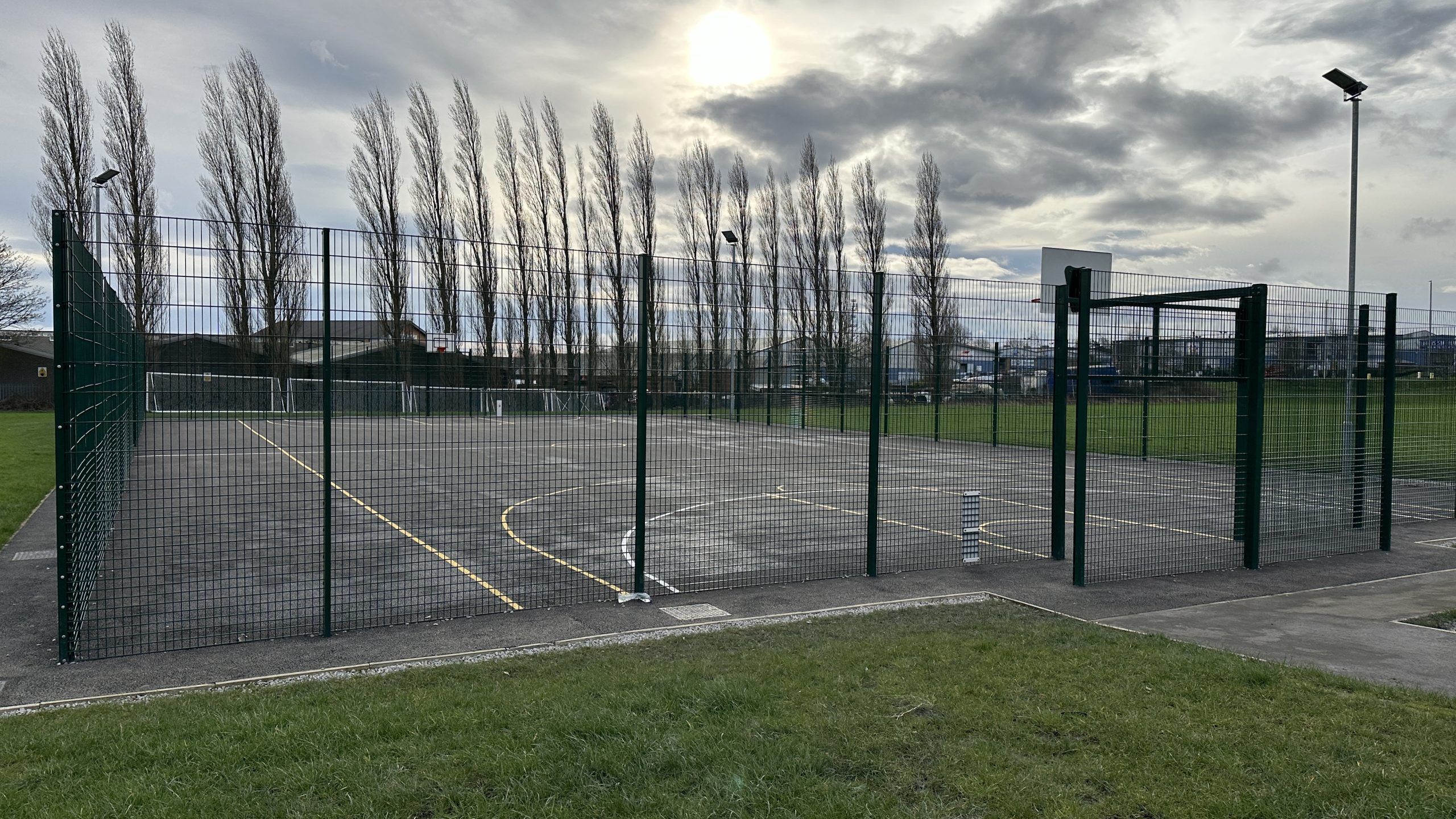 A multi-use games area with a basket ball and football markings, surrounding by high green fencing.